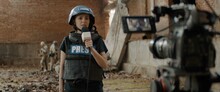 Behind The Scenes Of Female War Journalist Correspondent Wearing Bulletproof Vest And Helmet Reporting Live Near Destroyed Building, Military Personnel In The Background