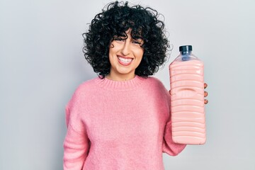 Sticker - Young middle east woman holding detergent bottle looking positive and happy standing and smiling with a confident smile showing teeth