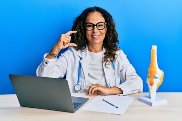 Poster - Beautiful middle age woman doctor at orthopedic clinic smiling and confident gesturing with hand doing small size sign with fingers looking and the camera. measure concept.