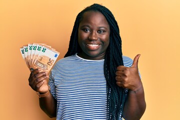 Sticker - Young black woman with braids holding bunch of 50 euro banknotes smiling happy and positive, thumb up doing excellent and approval sign