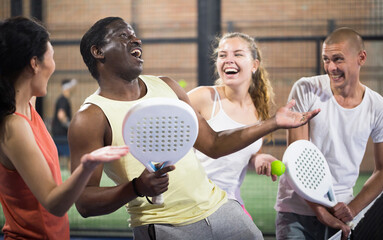 Multiethnic group of cheerful paddle tennis players standing on closed court after match having friendly chat. Focus on emotional expressive African American man