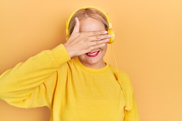 Canvas Print - Young blonde woman listening to music using headphones smiling and laughing with hand on face covering eyes for surprise. blind concept.