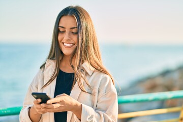 Wall Mural - Young hispanic girl smiling happy using smartphone at the promenade.