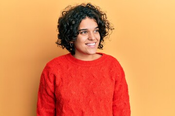 Young hispanic woman with curly hair wearing casual winter sweater looking away to side with smile on face, natural expression. laughing confident.