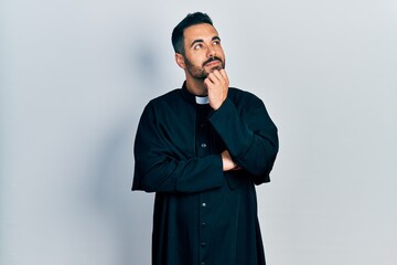 Wall Mural - Handsome hispanic man with beard wearing catholic priest robe with hand on chin thinking about question, pensive expression. smiling and thoughtful face. doubt concept.