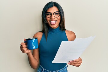 Wall Mural - Young latin transsexual transgender woman drinking cup of coffee and reading paper sticking tongue out happy with funny expression.