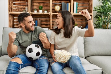 Wall Mural - Young latin couple watching soccer match eating porpcorn at home.