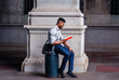 Wearing a gray shirt, blue jeans, black cloth shoes, carrying a shoulder leather bag, a young black man is sitting on a metal pillar on an old street in New York City, relaxing, reading a red book..