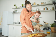 Waist Up Portrait Of Busy Mother Holding Baby Looking At Camera While Cooking In Kitchen, Copy Space
