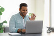 © kucherav - Friendly young Indian man using laptop for video communication with employees. Male freelancer in casual clothes waving hand in front of webcam greeting online participants