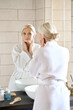 © insta_photos - Attractive beautiful mid age adult 50 years old blonde woman standing in bathroom of SPA hotel touching face, looking at reflection in mirror doing daily morning beauty routine. Skin care concept.