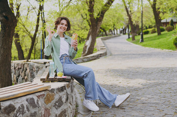 Full length young student woman in green jacket jeans sit on bench in spring park outdoors rest eat fast food hamburger have breakfast show okay ok gesture People unhealthy urban lifestyle concept