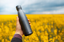 Close-up Of Male Hand Holding A Dark Grey, Steel Reusable Thermo Water Bottle On Background Of Blurred Rapeseed Field.