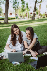 Two female students are sitting in the park on the grass with books and laptops, studying and preparing for exams. Distance education. Soft selective focus.