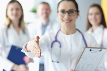 Woman doctor stretches out her hand for greetings against background of medical colleagues