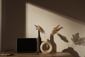 Wall Mural - Laptop computer with blank screen on table with pampas grass bouquet in sunlight shadows on the wall. Aesthetic boho office workspace interior design. Sunlight shade reflections