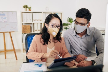 Wall Mural - Serious business colleagues in protective masks sitting at office desk sitting at office desk and discussing chart on screen of digital tablet