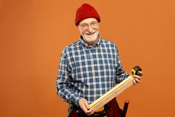 Successful professional attractive elderly male construction worker wearing overalls and belt kit of instruments posing in studio, smiling happily, measuring length of wooden plank using tool