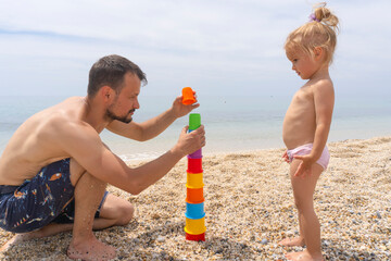 young caucasian man with little child play with colorful toys at sandy beach near the sea, summer ti