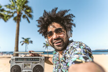 Young Latin Man Having Fun Taking Selfie With Mobile Smartphone While Listening Music With Headphones And Boombox On The Beach During Summer Vacations