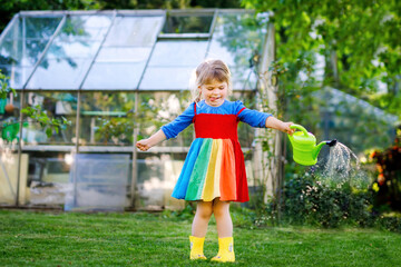 Wall Mural - Beautiful little toddler girl in yellow rubber boots and colorful dress watering spring flowers with kids water can. Happy child helping in family garden, outdoors.