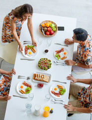 Top, aerial view Asian beautiful housewife in a colorful dress serving delicious tasty breakfast to family members at clean white table in comfortable dining room at home on bright refresh morning.