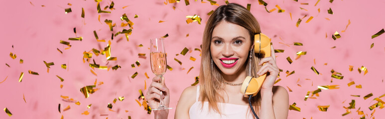 Wall Mural - Smiling woman holding champagne and talking on telephone near confetti on pink background, banner.