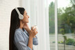 © New Africa - Young woman with cup of drink listening to music near window at home, space for text