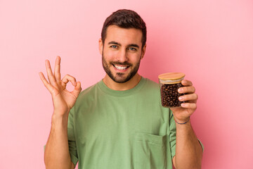 Wall Mural - Young caucasian man holding a coffee bottle isolated on pink background cheerful and confident showing ok gesture.