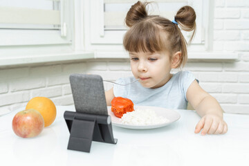 Little girl is having breakfast while watching the movie on the tablet