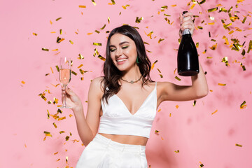 Wall Mural - Pretty brunette woman holding bottle and glass of champagne under golden confetti on pink background.