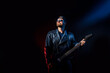 © zamuruev - Brutal bearded Heavy metal musician in leather jacket, cap and sunglasses is playing electrical guitar. Shot in a studio on dark background