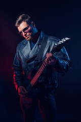 Brutal bearded Heavy metal musician in leather jacket, cap and sunglasses is playing electrical guitar. Shot in a studio on dark background