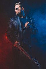 Brutal bearded Heavy metal musician in leather jacket, cap and sunglasses is playing electrical guitar. Shot in a studio on dark background