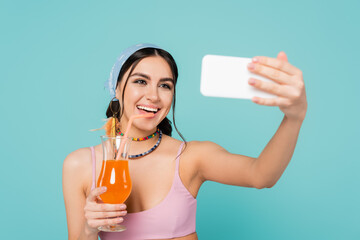 Wall Mural - Positive woman with cocktail taking selfie on blurred smartphone isolated on blue.