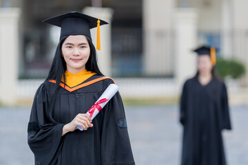 Wall Mural - A young beautiful Asian woman university graduate in graduation gown and mortarboard holds a degree certificate stands in front of the university building after participating in college commencement