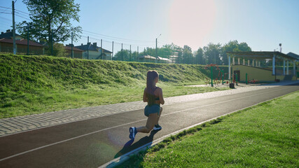 Back view of fit unrecognizable girl training legs, walking at stadium. Sexy, muscular woman warming up, practicing lunges outdoors in sunny summer morning. Sport, active lifestyle concept.