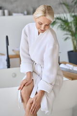 Wall Mural - Happy smiling gorgeous senior lady in bath robe at spa center sitting on bathtube touching legs. Advertising of bodycare spa procedures antiage recreation skin care salon concept. Vertical shot.