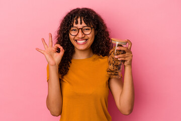 Young mixed race woman holding a cookies jar isolated on pink background cheerful and confident showing ok gesture.