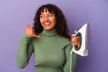 Wall Mural - Young mixed race woman holding an iron isolated on purple background showing a mobile phone call gesture with fingers.