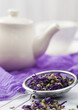 Strainer infuser with loose blue mallow flowers tea on purple cloth with ceramic teapot.