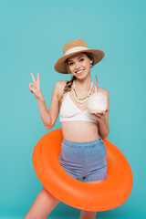 Wall Mural - Positive woman in swim ring holding cocktail in coconut and showing peace gesture isolated on blue.