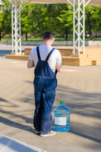 An Adult Man In Overalls Delivers Drinking Water To The Shops. A Worker Stands With A Barrel Of Drinking Water.