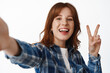 © Cookie Studio - Cheerful ginger girl student taking selfie with peace v-sign, smiling happy, posing for photo against isolated white background, standing in plaid shirt