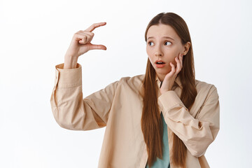 Wall Mural - Girl looks troubled at small tiny size, showing little thing and staring worried at product on copy space, standing against white background