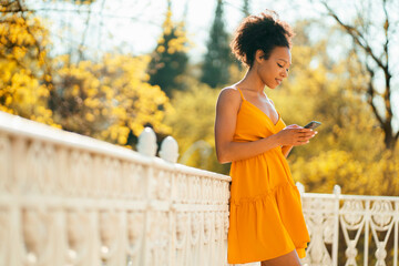 Wall Mural - Prints a message to the mail in the phone in the messenger. A beautiful female student of avro appearance in a summer dress of yellow gold color walks in a park in the center of the city.