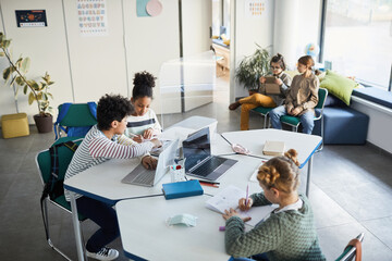 Wall Mural - High angle view at diverse group of children working together at desk in classroom with protective glass separations, copy space