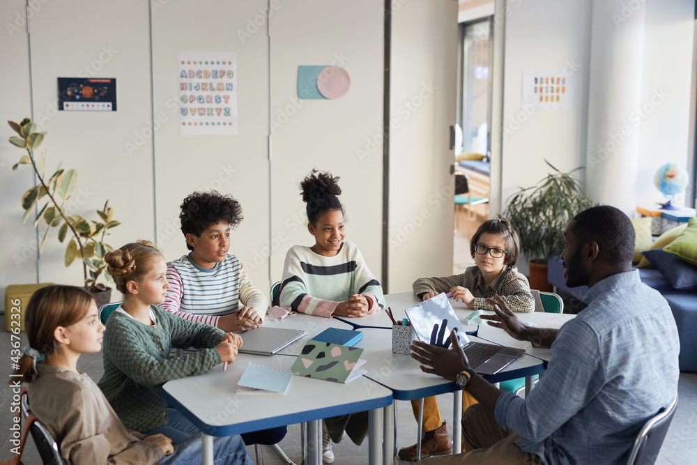 Diverse group of children sitting at table with male teacher in modern ...