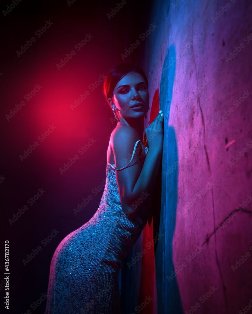 Sexy attractive young adult woman vamp in silver tight cocktail dress is standing at concrete wall in neon light, teasing over dark blue background. Beautiful women and sexual outfit concept - obrazy, fototapety, plakaty 