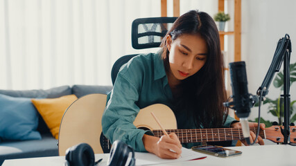 Wall Mural - Happy asia woman songwriter play acoustic guitar listen song from smartphone think and write notes lyrics song in paper sit in living room at home studio. Music production at home concept.